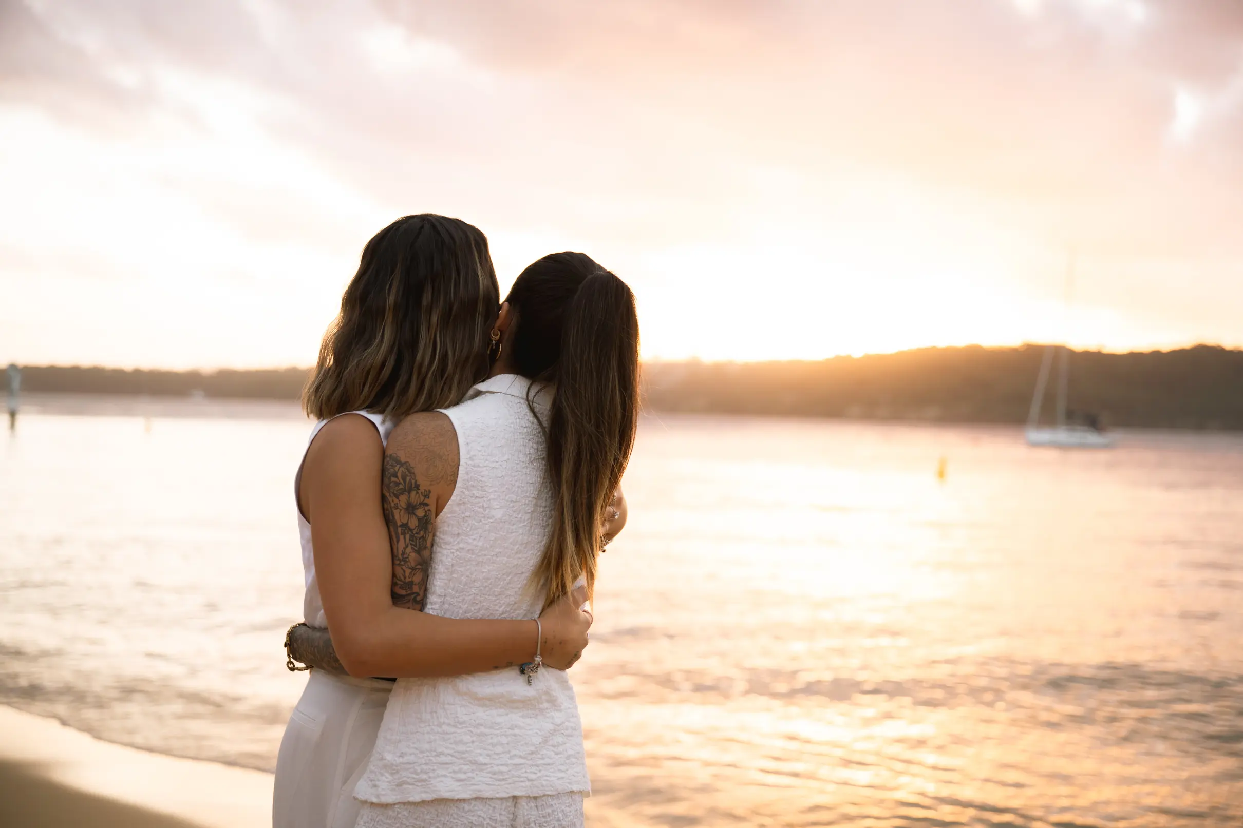 Two women embracing, backs to camera, looking out at the sunset water