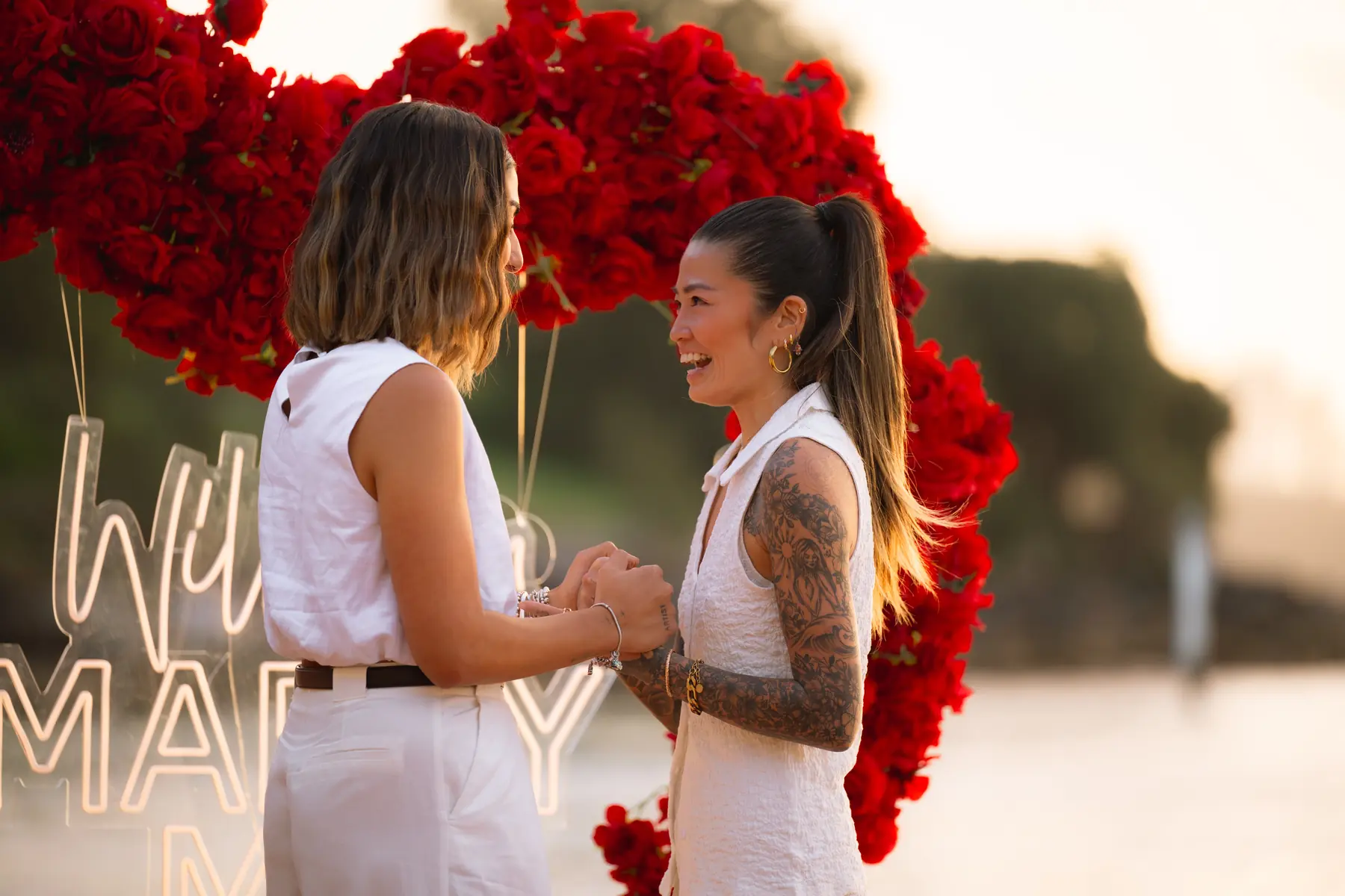 Two women holding hands and smiling in front of a red rose arch and Will You Marry Me neon sign