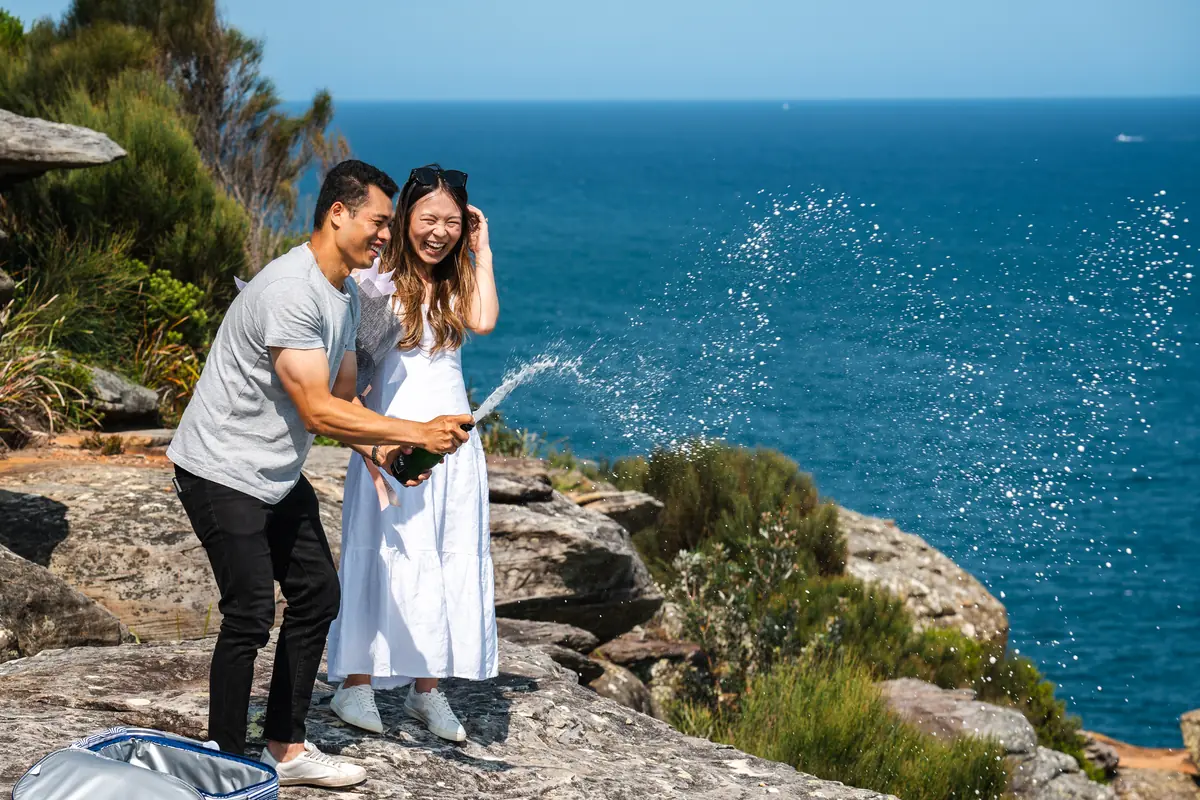 Newly engaged couple laughing as champagne sprays over a coastal clifftop