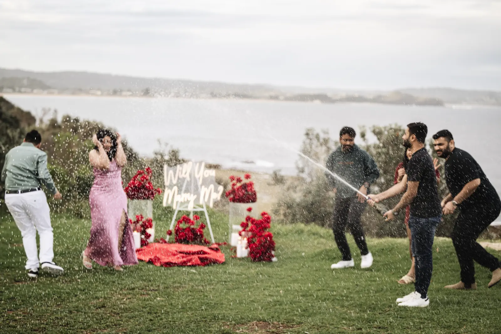 Friends spraying champagne at a headland proposal set-up with red roses and a neon sign